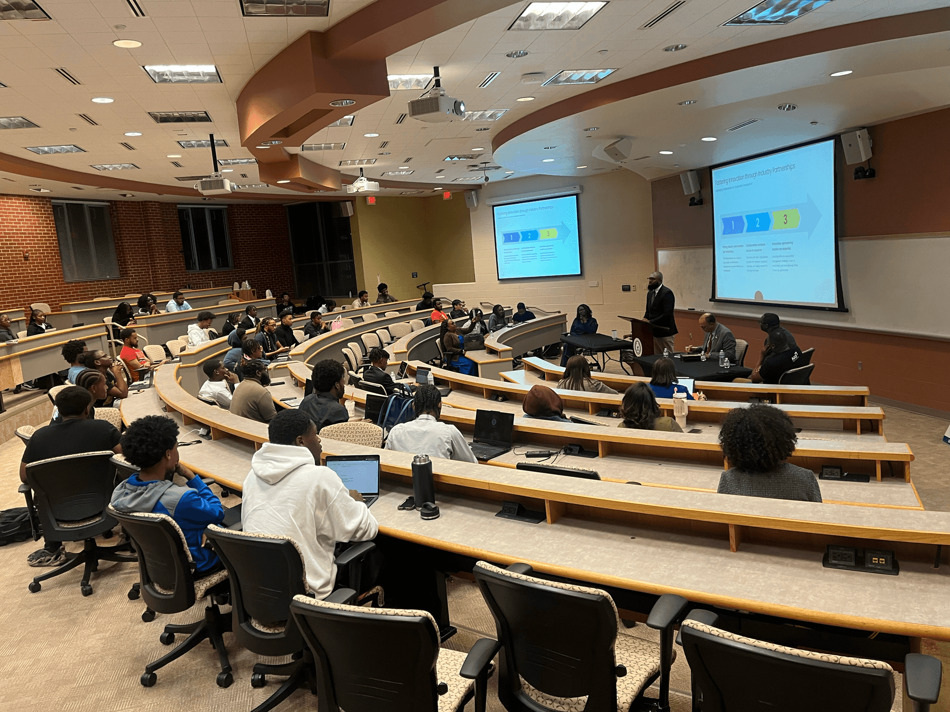 Lecture hall with students attending a presentation, speaker standing near a screen displaying slides.