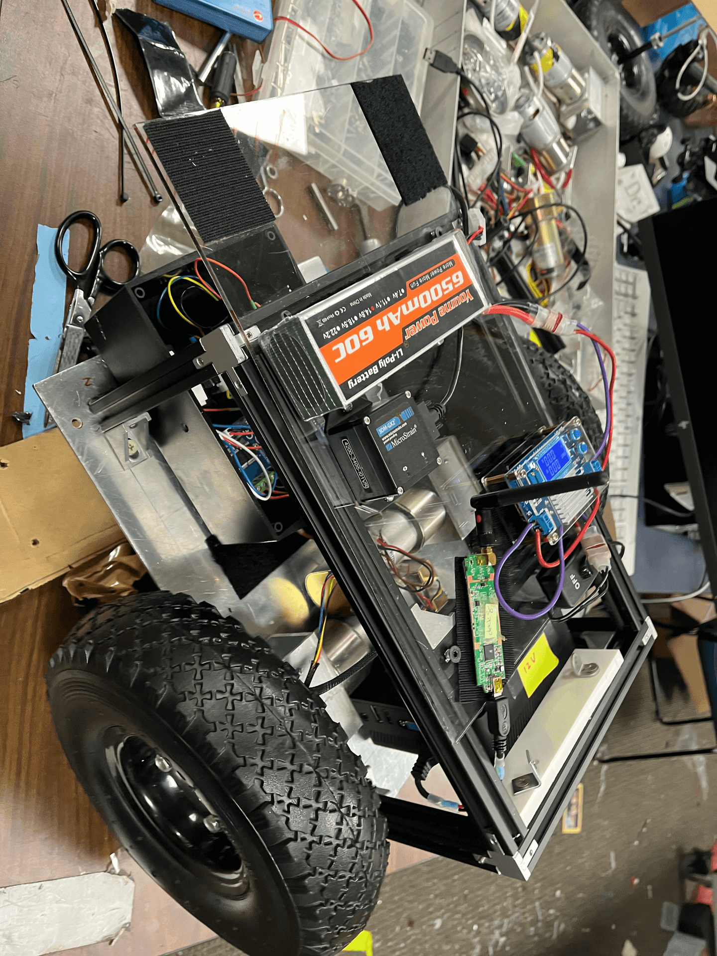 DIY robot vehicle with electronics and wires on a workbench, featuring large wheels and battery pack.